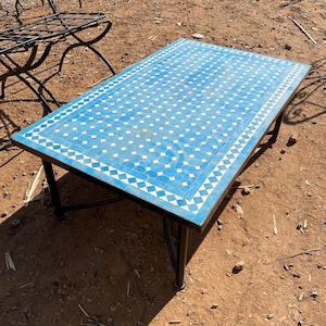 Peut inclure: Table rectangulaire d'extérieur avec un dessus en mosaïque de carreaux bleus et blancs. La table a un cadre et des pieds en métal noir. Le motif de la mosaïque présente un motif géométrique répétitif. La table mesure environ 152 cm de long et 76 cm de large.