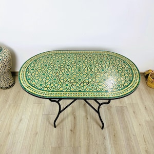 May include: Oval-shaped table with a green and yellow mosaic tile top and a black metal frame. The table is on a wooden floor.