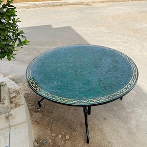 Könnte beinhalten: Ein runder, grüner Mosaiktisch mit einem schwarzen Metallgestell. Die Tischplatte besteht aus kleinen, grünen Fliesen mit einem goldenen Rand.