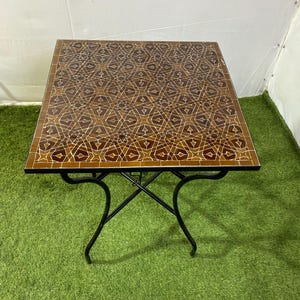 Peut inclure: Table carrée en mosaïque avec un motif géométrique marron et beige. La table a un piètement en fer forgé noir. La table mesure environ 61 cm de côté.