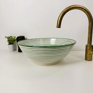 May include: A white ceramic bowl with green stripes. The bowl is round and has a wide rim. It is sitting on a white surface. A gold tap is in the background.