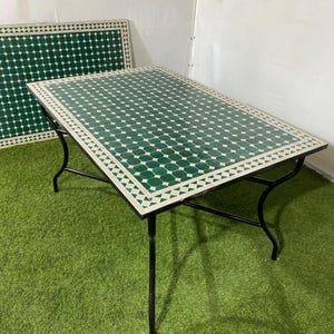 May include: A rectangular outdoor table with a green and white tiled top and black metal legs. The tabletop features a repeating pattern of green diamonds and white triangles around the edges. A matching tile panel is visible in the background.
