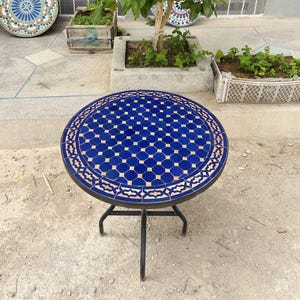 Könnte beinhalten: Ein runder, blau-weißer Tisch mit Fliesen und einem schwarzen Metallgestell. Die Tischplatte hat ein geometrisches Muster mit einem rosa-weißen Rand.
