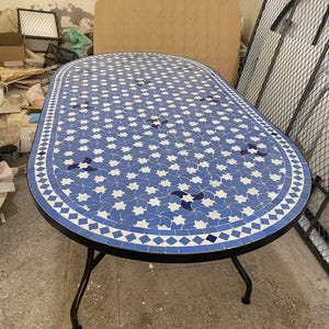 May include: Oval mosaic tile table with a blue and white star pattern. The table has a black metal frame and legs. The table is approximately 152 cm long and 76 cm wide.