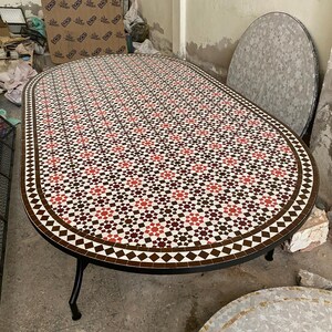 May include: An oval mosaic tile table with a black metal frame. The tabletop features a repeating pattern of red, brown, and white tiles. The table is approximately 152 cm long and 76 cm wide.