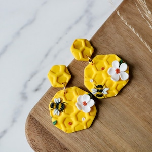 Könnte beinhalten: Ein Paar gelbe Honigwaben-Ohrringe mit weißen Blumen und Bienen. Die Ohrringe sind aus Polymer Clay gefertigt und haben einen goldfarbenen Haken.