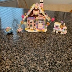 Può includere: Una casetta di pan di zenzero rosa e marrone con decorazioni di caramelle, un'auto giocattolo blu con caramelle in cima, un coniglietto rosa e bianco con un lecca-lecca e un trenino giocattolo rosa e blu con decorazioni di caramelle.