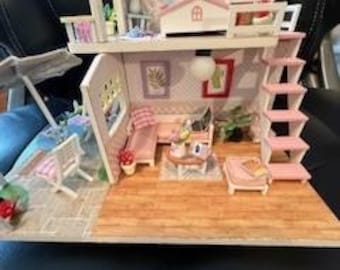 Puede incluir: Una casa de muñecas en miniatura con un exterior rosa y blanco, que incluye un balcón, una escalera y una sala de estar con muebles. La casa de muñecas está decorada con motivos florales y tiene un pequeño patio con sillas y una sombrilla.