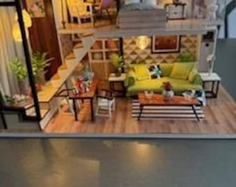 May include: Miniature two-story dollhouse with a living room and bedroom. The living room features a green sofa, coffee table, and rug. The bedroom has a bed and nightstand. The house includes a staircase, bookshelves, and decorative lighting.
