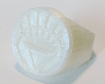 Può includere: Un anello bianco con una faccia circolare con un triangolo all'interno di un disegno a raggiera. L'anello ha una fascia spessa e una superficie liscia e arrotondata. L'anello è realizzato in un materiale di colore chiaro.