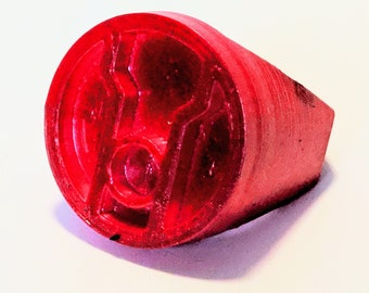 Può includere: Un anello rosso con un design geometrico su sfondo bianco. L'anello è di un colore rosso vivo e sembra essere fatto di un materiale liscio. Il design sull'anello è una serie di forme angolari.