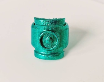 Può includere: Un anello verde turchese con un design circolare in rilievo al centro. L'anello è fatto di un materiale scintillante.