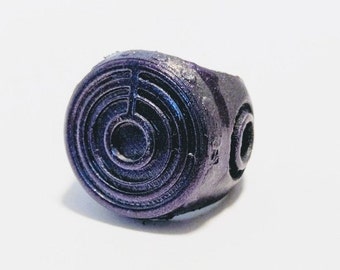 Può includere: Un anello viola con un design circolare che presenta cerchi concentrici e un cerchio più piccolo al centro. L'anello è realizzato in un materiale liscio e lucido.