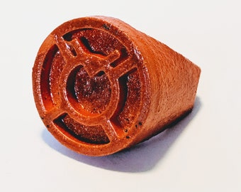 Può includere: Un anello marrone con un simbolo arancione in rilievo al centro. Il simbolo è un cerchio con un cerchio più piccolo all'interno e una linea che attraversa il centro.
