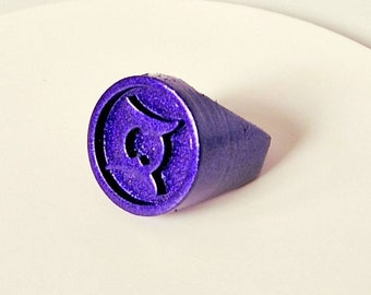 Può includere: Un anello viola con un design stilizzato sulla parte superiore. L'anello è realizzato in un materiale liscio e lucido.