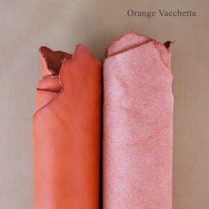 May include: Two rolled pieces of leather in shades of orange and pink. The orange leather is smooth, while the pink leather has a suede-like texture. The text "Orange Vacchetta" is at the top of the image.