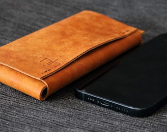 Puede incluir: Una cartera de cuero marrón claro con solapa plegada y detalles cosidos. Junto a la cartera hay un smartphone negro. La cartera tiene un diseño minimalista y parece estar hecha de cuero de alta calidad.
