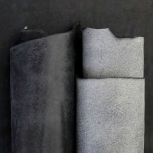 May include: Two rectangular pieces of leather material are displayed side by side. The piece on the left is a deep, solid black with a smooth, matte finish. The piece on the right is a lighter, textured gray with a visible grain and rough edges.