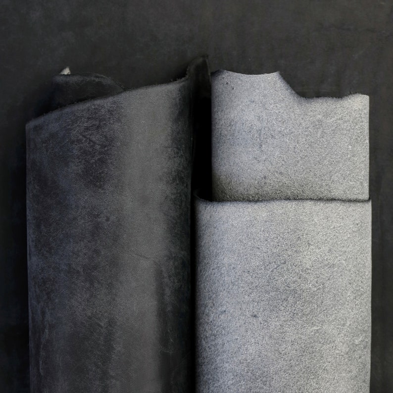 May include: Two rectangular pieces of leather material are displayed side by side. The piece on the left is a deep, solid black with a smooth, matte finish. The piece on the right is a lighter, textured gray with a visible grain and rough edges.