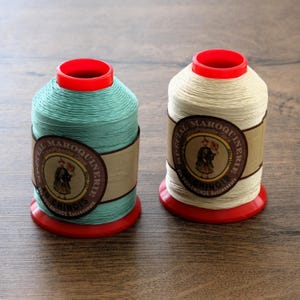 May include: Two spools of thread with bright red plastic bases and tops. One spool is light mint green, and the other is off-white. Each features a tan paper label with the text Special Maroquinerie, Au Chinois, and an illustration of a person.