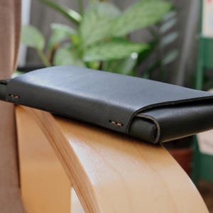 Puede incluir: Una cartera de cuero verde oscuro con solapa curva. La cartera rectangular tiene costuras visibles y una textura suave. Descansa sobre una superficie de madera clara.