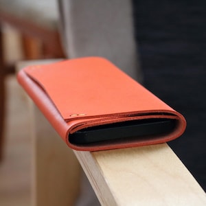 Puede incluir: Una funda de teléfono de cuero naranja brillante con una solapa doblada. La funda está sobre una superficie de madera.