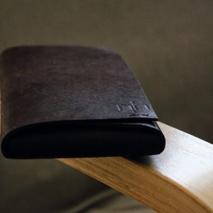 May include: A dark brown leather wallet with a stitched seam and a small embossed logo. The wallet is resting on a light brown wooden surface.