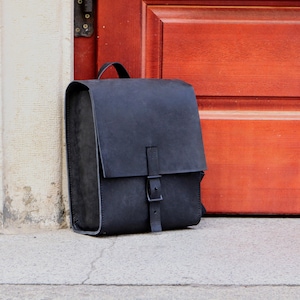 May include: A black leather backpack with a rectangular shape and a top handle. The bag features a flap closure with a buckle and a strap. The backpack is leaning against a wall and a red door.