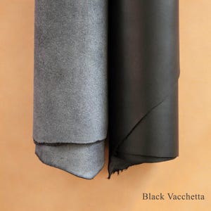 May include: Two rolls of leather, one gray suede and one black, with the text "Black Vacchetta" at the bottom. The gray leather has a soft, textured surface, while the black leather is smooth and shiny.