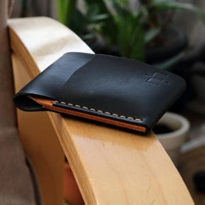 Peut inclure: Un portefeuille en cuir noir au design minimaliste. Le portefeuille présente une construction pliée en deux avec des coutures visibles en fil marron clair contrastant. Un logo subtil en relief est visible sur le devant. Le portefeuille mesure environ 10 cm de large.