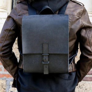 May include: A dark gray leather backpack with a rectangular design. The backpack features a flap closure secured by a matching strap and a metal buckle. The bag has a top handle and adjustable shoulder straps.