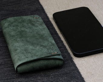 May include: A dark green leather phone case with a folded design, next to a black smartphone. The case has a textured appearance and is stitched with orange thread. The phone is black and has a sleek, modern design.