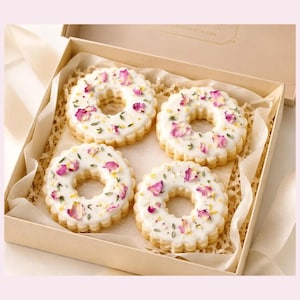 May include: Four round, scalloped-edge cookies arranged in a square formation inside a tan cardboard gift box. Each cookie is coated in a smooth white icing and topped with scattered dried pink rose petals and small green herb sprigs.