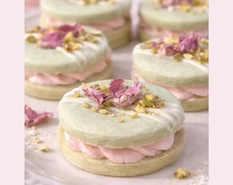 Könnte beinhalten: Fünf runde Sandwich-Kekse bestehend aus hellgrünen runden Keksen mit einer Füllung aus hellrosa Creme. Die Oberseiten sind mit weißem Zuckerguss, gehackten gelben Nüssen und getrockneten rosa Rosenblättern verziert und auf einem Teller angerichtet.