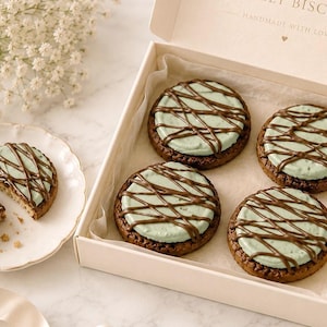 May include: A box containing four round chocolate cookies topped with light green icing and a dark chocolate drizzle. A small plate holds one additional cookie broken into two pieces. The box lid features the text "HANDMADE WITH LOVE".