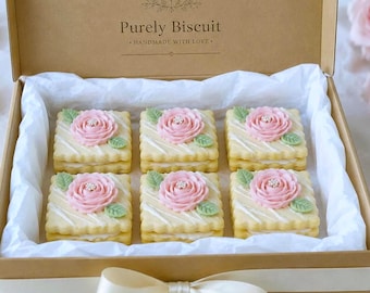 Può includere: Una scatola regalo in cartone con l'etichetta Purely Biscuit Handmade With Love contenente sei biscotti quadrati farciti. Ogni biscotto presenta una rosa di glassa rosa, una piccola foglia verde e una decorazione bianca su un bordo smerlato.