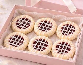 May include: Six round, light-colored cookies with a dark red jam center and a white icing lattice pattern on top, arranged inside a light pink gift box lined with white parchment paper. The cookies have a scalloped edge design.