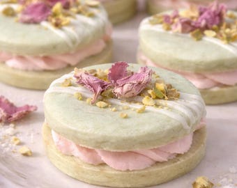 May include: Round sandwich cookies featuring pale green circular biscuits filled with light pink cream. The top of each cookie is decorated with a white icing drizzle, crushed green pistachio nuts, and dried pink rose petals.