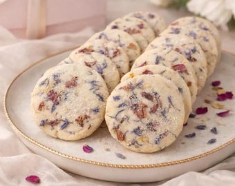 May include: A plate of round cookies, each adorned with colorful flower petals and a dusting of sugar. The cookies are arranged on a white plate with a gold rim, alongside a pink gift box with a ribbon.