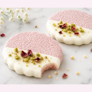 May include: Two round pink cookies with a textured surface, each partially dipped in smooth white icing. The iced sections are topped with small green pistachio pieces and dried red rose petals. One cookie has a visible bite mark on the edge.