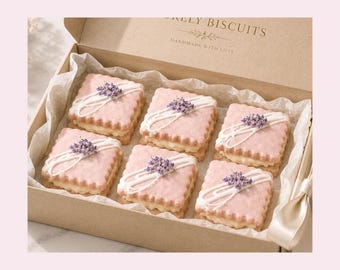 Könnte beinhalten: Eine Geschenkschachtel aus Karton mit sechs quadratischen Sandwich-Keksen mit rosa gewelltem Rand, einer Cremefüllung, weißen Glasurstreifen und kleinen violetten Lavendelzweigen. Der Deckel trägt den Text "RELY BISCUITS HANDMADE WITH LOVE".