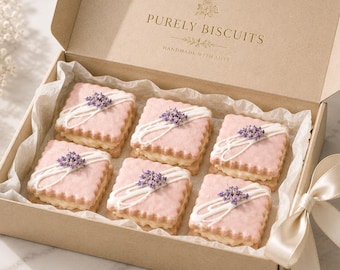 May include: A cardboard gift box containing six square sandwich biscuits with pink scalloped edges, a cream filling, white icing drizzles, and small purple lavender sprigs on top. The box lid features the gold text PURELY BISCUITS HANDMADE WITH LOVE.