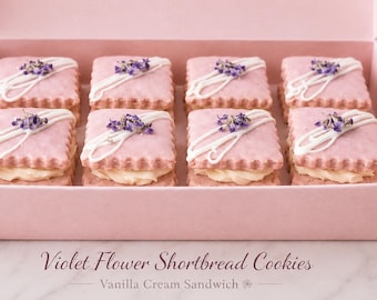 May include: A pink box containing eight square shortbread sandwich cookies with pink icing, white icing drizzles, and small purple flowers on top. The cookies are filled with a pale yellow cream. Text below reads Violet Flower Shortbread Cookies Vanilla Cream Sandwich.