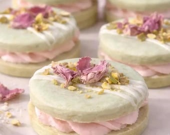 Może przedstawiać: Okrągłe ciasteczka typu sandwich z jasnozielonymi krążkami, jasnoróżowym kremem, białym lukrem, posiekanymi pistacjami i suszonymi płatkami róż. Tekst na dole brzmi Gluten free/vegan option.