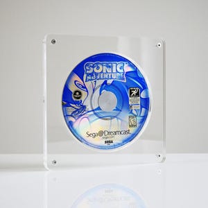 Puede incluir: Un disco del juego Sonic Adventure para Dreamcast exhibido en un marco de acrílico transparente. El disco presenta un diseño azul y blanco con el título del juego y la marca Sega Dreamcast. El marco cuadrado tiene cabezas de tornillo visibles.
