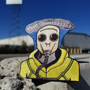 May include: A yellow and blue embroidered patch of a person wearing a gas mask with the text "What Chemtrails 1s?" above the person's head.
