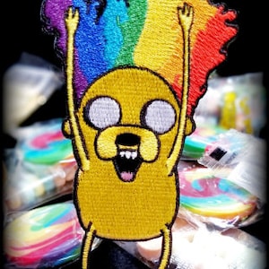May include: Embroidered patch of a yellow cartoon dog with a rainbow afro, the dog is smiling and has its arms raised in the air.
