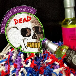 May include: A white skull with a green and purple embroidered patch that says "I'll sleep when I'm dead". The skull is wearing gold sunglasses and has a gold tooth. The skull is sitting on a bed of colorful shredded paper.