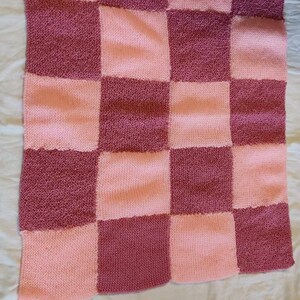 Può includere: Una copertina per neonati in maglia rosa e malva con un motivo a scacchi.