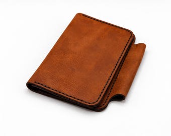 May include: A rectangular brown leather wallet featuring visible dark stitching along the edges and a folded flap design on the right side. The leather has a smooth, matte texture with rounded corners and a consistent tan color throughout.